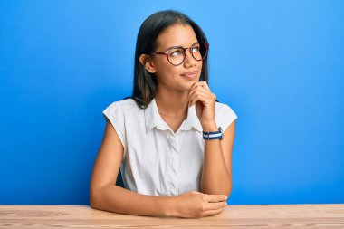 Güzel İspanyol kadın, gündelik giysiler giymiş, elleri çenesinde masada oturmuş soru sormayı düşünüyor, düşünceli yüz ifadesi. Gülümseyen ve düşünceli bir yüz. şüphe kavramı. 