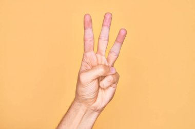 İzole edilmiş sarı arka planda parmaklarını gösteren beyaz bir gencin eli üç parmağı gösteriyor.