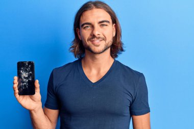 Kırık akıllı telefon tutan genç yakışıklı adam kırık ekranı gösteriyor. Pozitif ve mutlu görünüyor. Kendine güvenen bir gülümsemeyle dişlerini gösteriyor. 