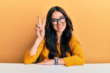 Güzel, esmer, gözlüklü, sıradan kıyafetler giyen genç bir kadın masanın üzerinde oturuyor ve parmaklarını iki numarayı göstererek gülümsüyor. Kendinden emin ve mutlu.. 