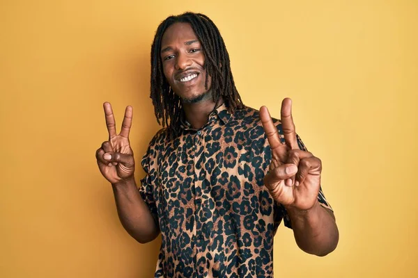 Saçları örgülü, leopar desenli hayvan desenli tişörtlü, parmaklarını zafer işareti yaparken gösteren kameraya bakan Afro-Amerikan bir adam. İki numara.. 