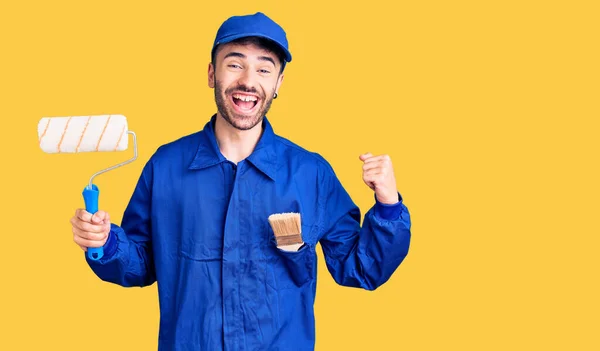 Joven Hombre Hispano Vistiendo Uniforme Pintor Sosteniendo Rodillo Gritando Orgulloso —  Fotos de Stock