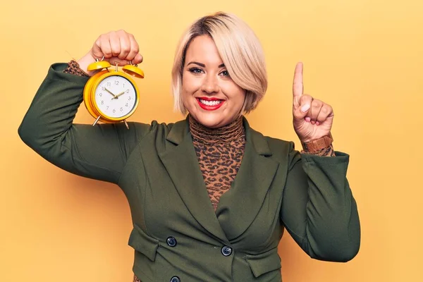 Young beautiful plus size blonde woman holding vintage alarm clock over yellow background smiling with an idea or question pointing finger with happy face, number one