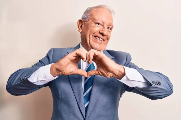 Senior handsome grey-haired businessman wearing elegant suit over white background smiling in love doing heart symbol shape with hands. Romantic concept.