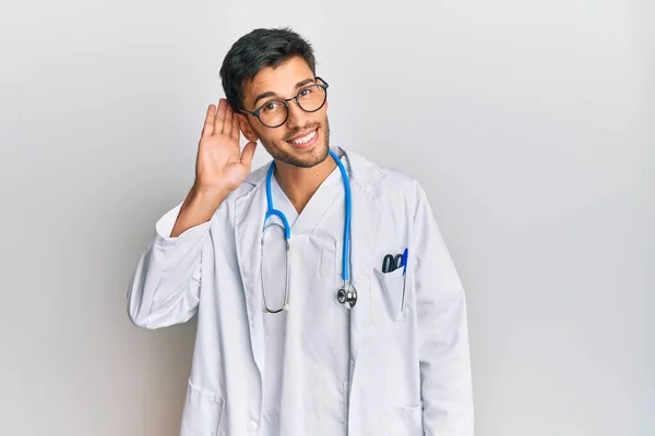 医師の制服を着た若いハンサムな男と聴診器は噂やゴシップに耳を傾ける耳の上に手で笑みを浮かべて 聴覚障害の概念 — ストック写真