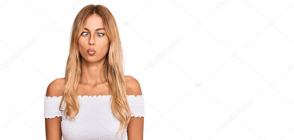 Beautiful blonde young woman wearing casual white tshirt making fish face with lips, crazy and comical gesture. funny expression. 