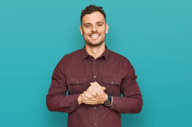 Latin asıllı genç adam ellerinin birleştiği günlük kıyafetler giyiyor ve gülümseyerek gevşemiş ve neşeli gülümsüyor. Başarı ve iyimserlik 