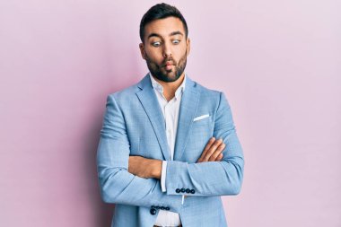 Young hispanic businessman wearing suit with arms crossed gesture making fish face with mouth and squinting eyes, crazy and comical. 