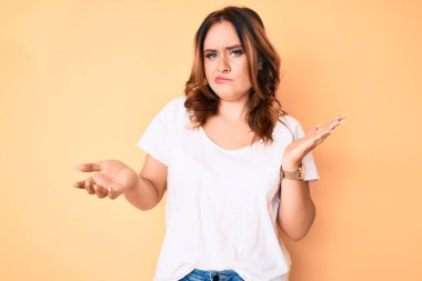 Beyaz günlük beyaz tişört giyen, kafası karışmış, kolları ve elleri havada genç, güzel beyaz bir kadın. şüphe kavramı. 