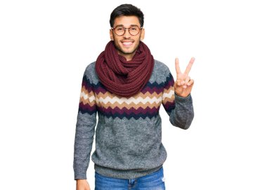 Kış süveteri giymiş genç yakışıklı bir adam. Gülümsüyor ve kameraya göz kırpıyor. Zafer işareti yapıyor. İki numara.. 