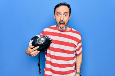 Orta yaşlı yakışıklı motosikletçi, moto kaskını mavi arka planda tutuyor. Şaşırmak için ağzı açık kalmış, korkmuş ve hayrete düşmüş. İnanmayan yüz.