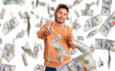 Kış süveteri ve yün şapka giyen yakışıklı Latin Amerikalı genç adam kameraya bakıp zafer işareti yapıyor. İki numara..