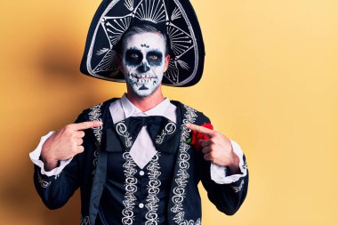 Young man wearing mexican day of the dead costume over yellow looking confident with smile on face, pointing oneself with fingers proud and happy. 