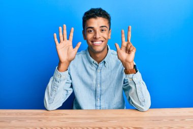 Genç, yakışıklı, Afro-Amerikalı, gündelik giysiler giymiş masada oturuyor ve 8 numaralı parmaklarıyla işaret ediyor, kendinden emin ve mutlu gülümsüyor.. 