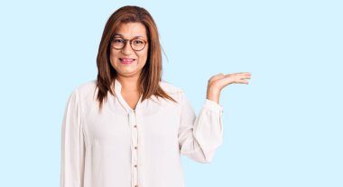 Orta yaşlı, gündelik kıyafetler ve gözlük takan, neşeli bir şekilde gülümseyen ve avuç içi kameraya bakan Latin bir kadın.. 