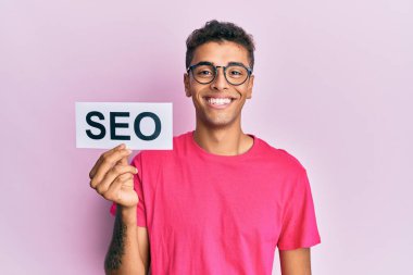 Genç, yakışıklı, Afro-Amerikan bir adam elinde seo-mesaj kağıdı tutuyor. Pozitif ve mutlu görünüyor. Dişlerini gösteren bir gülümsemeyle ayakta duruyor. 