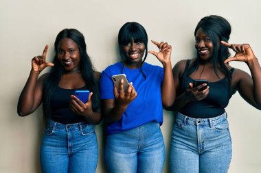 Üç Afro-Amerikalı genç arkadaş akıllı telefon kullanıyor gülümsüyor ve el hareketleriyle el hareketleriyle küçük boy işareti yapıyor ve kameraya bakıyor. ölçüm kavramı. 
