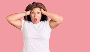 Orta yaşlı, beyaz tişört giyen, çılgın, elleri kafasında korkmuş, ağzı açık bir şekilde şok geçirmekten korkan Latin bir kadın. 