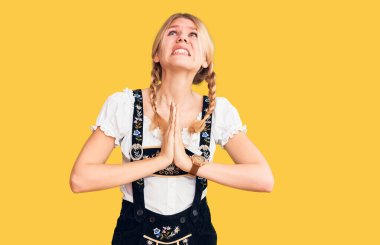 Oktoberfest elbisesi giyen genç, sarışın bir kadın elleri bağlı yalvarıyor ve dua ediyor. Yüzünde umut ifadesi var. Çok duygusal ve endişeli. Yalvarıyor. 