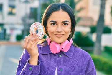 Genç İspanyol kadın şehirde kulaklık takıyor ve çörek yiyor..