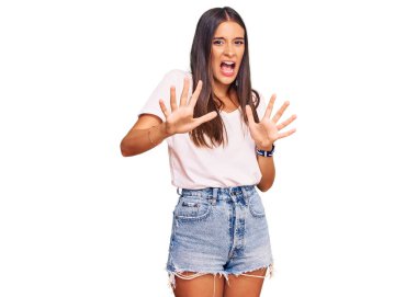 Young hispanic woman wearing casual white tshirt afraid and terrified with fear expression stop gesture with hands, shouting in shock. panic concept. 