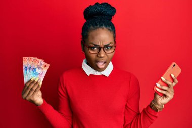 Genç Afro-Amerikalı kadın elinde İsviçre frangı banknotları olan akıllı telefon kullanıyor. Şüpheci ve alaycı görünüyor. 