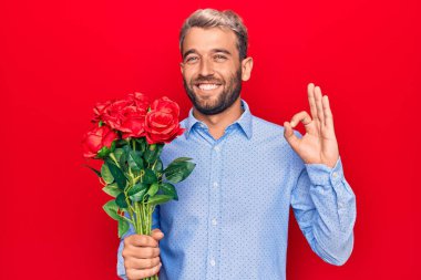 Genç, sarışın, romantik bir adam kırmızı arka planda güzel güller tutuyor. Tamam işareti yapıyor. Parmaklarıyla, gülümsüyor.