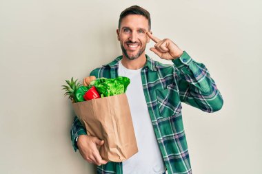 Handsome man with beard holding paper bag with groceries smiling pointing to head with one finger, great idea or thought, good memory 