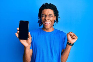 Young african american man holding smartphone showing blank screen screaming proud, celebrating victory and success very excited with raised arm 