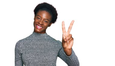 Genç Afro-Amerikan kızı gündelik giysiler giyip iki numaralı parmaklarıyla işaret ederken kendinden emin ve mutlu bir şekilde gülümsüyor.. 