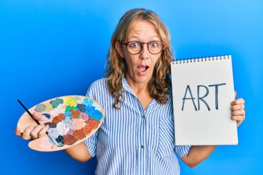 Orta yaşlı sarışın kadın elinde ressam paleti ve not defteri tutarken şaşkınlık ve şaşkınlık dolu bir ifade, korku ve heyecan dolu bir yüz ifadesi vardı.. 