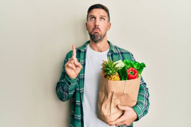 Handsome man with beard holding paper bag with groceries pointing up looking sad and upset, indicating direction with fingers, unhappy and depressed. 