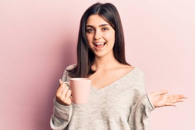 Genç, güzel bir kız bir kupa kahve içip başarıyı kutluyor mutlu bir gülümsemeyle ve kazanan bir ifadeyle elini kaldırarak. 