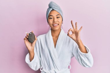 Duş havlusu şapkası ve bornoz giyen genç Çinli kadın sünger taşı tutuyor. Parmaklarıyla imza atıyor, gülümsüyor. 