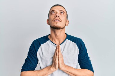 Hispanik genç adam gündelik giysiler giyip elleri bağlı yalvarıyor ve dua ediyor. Yüzünde umut ifadesi var. Çok duygusal ve endişeli. Yalvarıyor. 