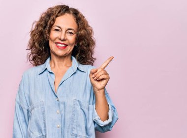 Orta yaşlı güzel bir kadın pembe arka planda dikilmiş sıradan bir kot gömlek giyiyor yüzünde kocaman bir gülümseme, eli kameraya bakıyor..