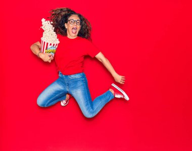 Orta yaşlı, güzel İspanyol bir kadın ağzı açık ve gözlük takıyor. İzole edilmiş kırmızı arka planda zıplayan patlamış mısır kovası.