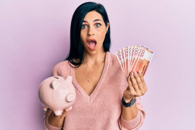 Elinde Norveç kronu banknotları ve domuz kumbarası olan beyaz bir kadın şaşkınlık ve şaşkınlık ifadesi, korku ve heyecan dolu bir yüzle korku ve şok içinde.. 