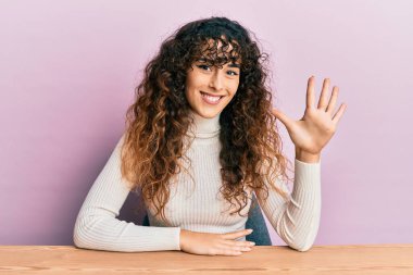 Latin kökenli genç bir kız, gündelik giysiler içinde masada oturuyor ve beş numaralı parmaklarıyla işaret ediyor, gülümsüyor ve mutlu.. 