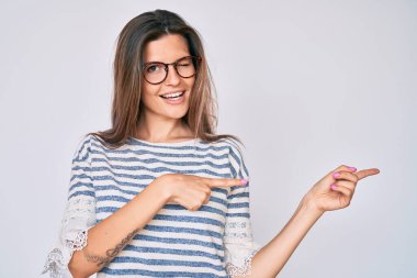 Güzel beyaz kadın, günlük kıyafetler ve gözlük takıyor gülümsüyor ve kameraya bakıyor. İki eliyle ve parmaklarıyla yandan işaret ediyor.. 