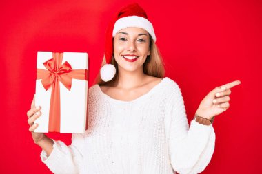 Genç esmer kadın Noel şapkası takıyor ve elinde bir hediye tutuyor mutlu bir şekilde gülümsüyor. 