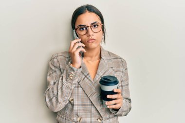 Genç esmer kadın akıllı telefon kullanıyor ve bir fincan kahve içiyor. Yüzünde ciddi bir ifade var. Basit ve doğal bir şekilde kameraya bakıyorsun.. 
