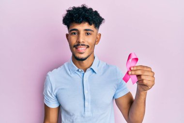 Genç Arap yakışıklı adam pembe kanser kurdelesi tutuyor. Pozitif ve mutlu görünüyor. Kendine güvenen bir gülümsemeyle dişlerini gösteriyor. 