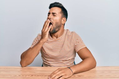 Masada oturan, gündelik kıyafetler giyen yakışıklı, esnemekten sıkılmış, ağzı eliyle kapatan İspanyol bir adam. Huzursuz ve uykulu.. 