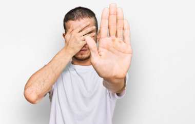 Latin kökenli genç adam, günlük beyaz tişörtler giyip elleriyle gözlerini kapatıyor ve üzgün ve korku ifadeleriyle jestleri durduruyor. utangaç ve olumsuz kavram. 