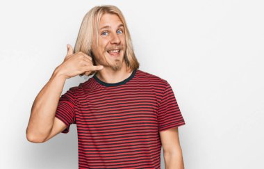 Caucasian man with blond long hair wearing casual striped t shirt smiling doing phone gesture with hand and fingers like talking on the telephone. communicating concepts. 