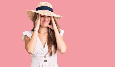 Genç, sarışın, yaz elbisesi giyen, şapkalı, baş ağrısı çeken çaresiz ve stresli bir kadın. Çünkü acı ve migren. Eller başının üstünde.. 