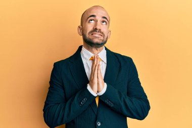 Genç İspanyol bir adam iş elbisesi ve kravat takıyor. Elleriyle yalvarıyor ve dua ediyor. Yüzünde umut ifadesi var. Yalvarıyor. 