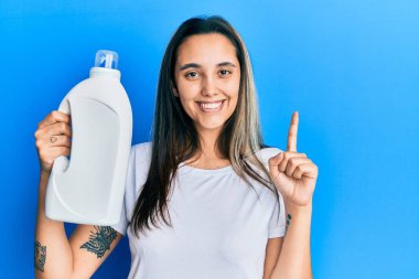 Elinde deterjan şişesi olan genç İspanyol bir kadın bir fikir veya soru ile gülümsüyor ve mutlu bir yüzle işaret ediyor. 