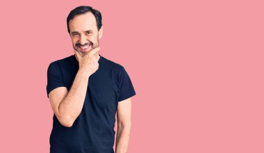 Orta yaşlı, sıradan tişörtler giyen yakışıklı bir adam kameraya kendinden emin bakıyor kollarını kavuşturmuş ve çenesini kaldırmış gülümsüyor. Olumlu düşünmek. 